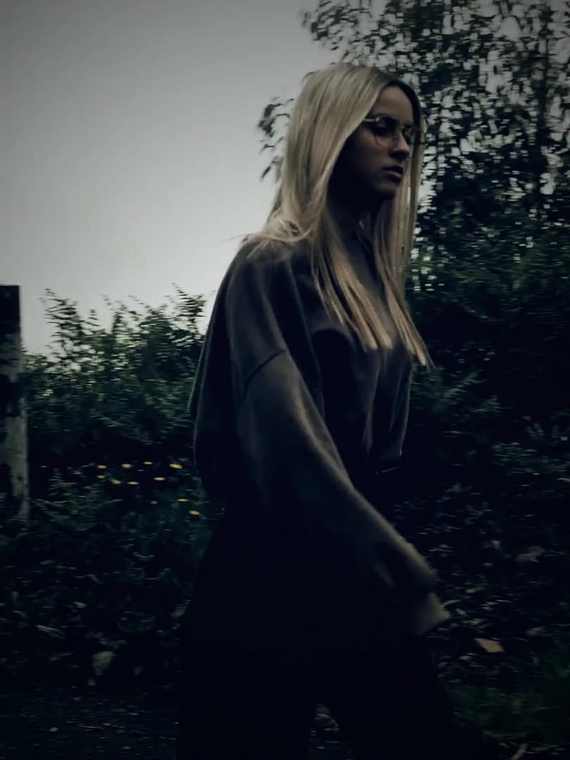 A woman in a long knit cardigan walking along a misty roadside.