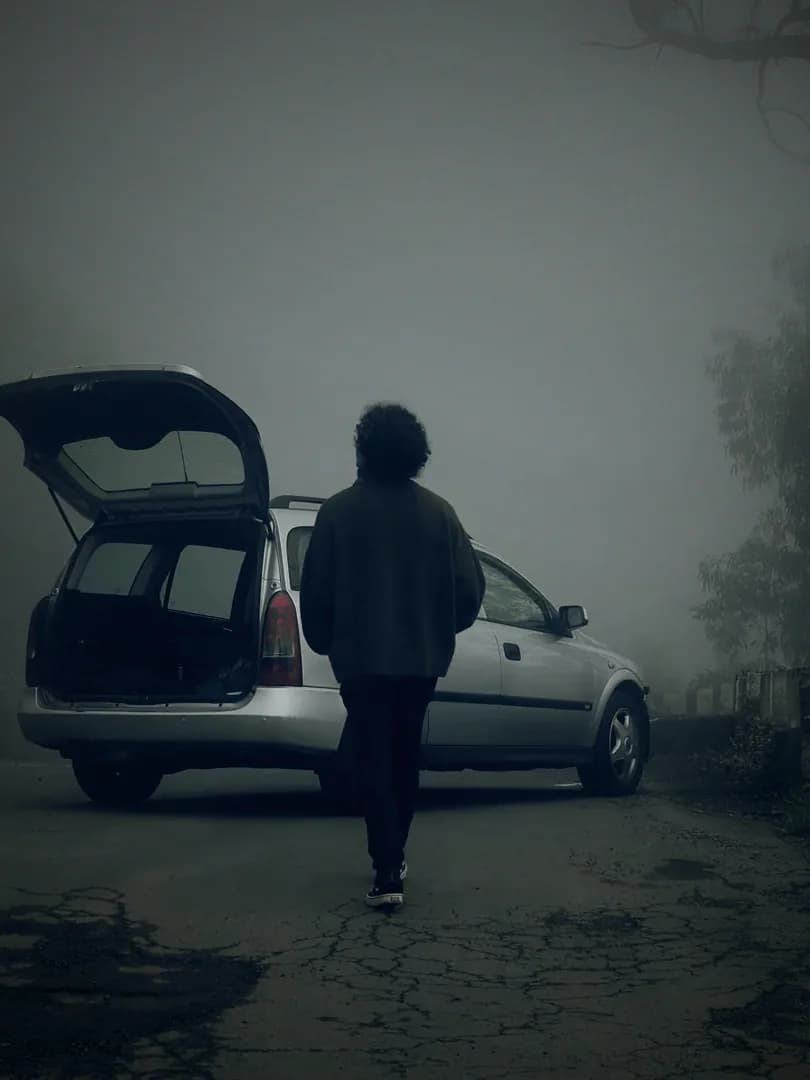 A figure walking back toward a parked car on a road lost in fog.