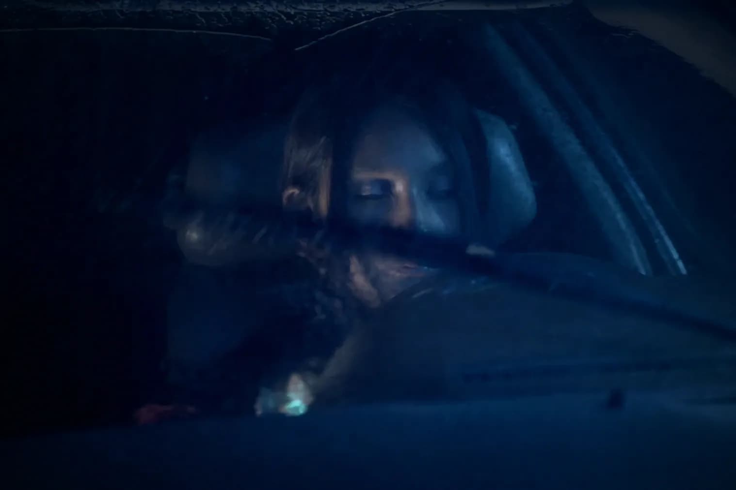 A woman's face seen through a rain-streaked car window in cold blue light.