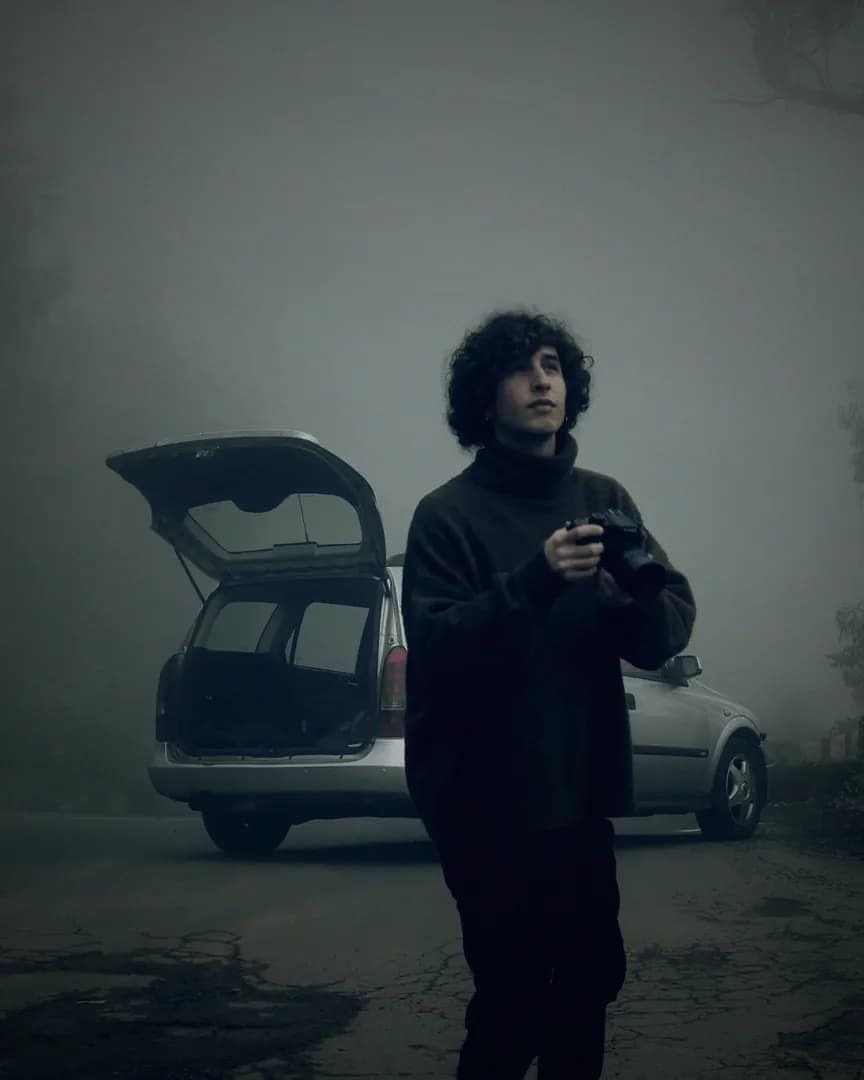 A young man with a camera standing on a foggy mountain road beside an estate car with its boot open.
