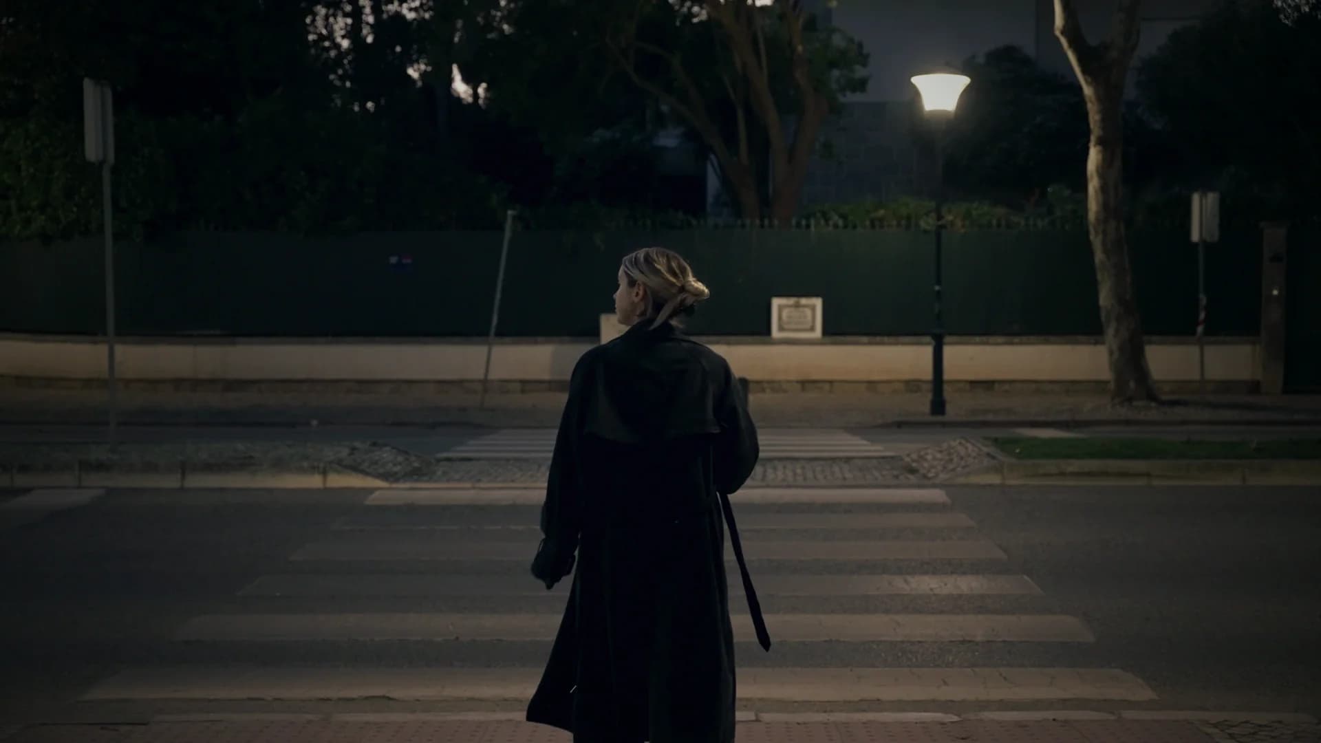 A woman in a long dark coat crossing an empty street under a streetlamp at nightfall.