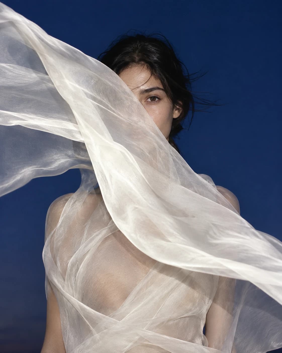 AI fashion editorial of a woman half hidden behind a sheer white silk veil blowing against a deep cobalt sky.
