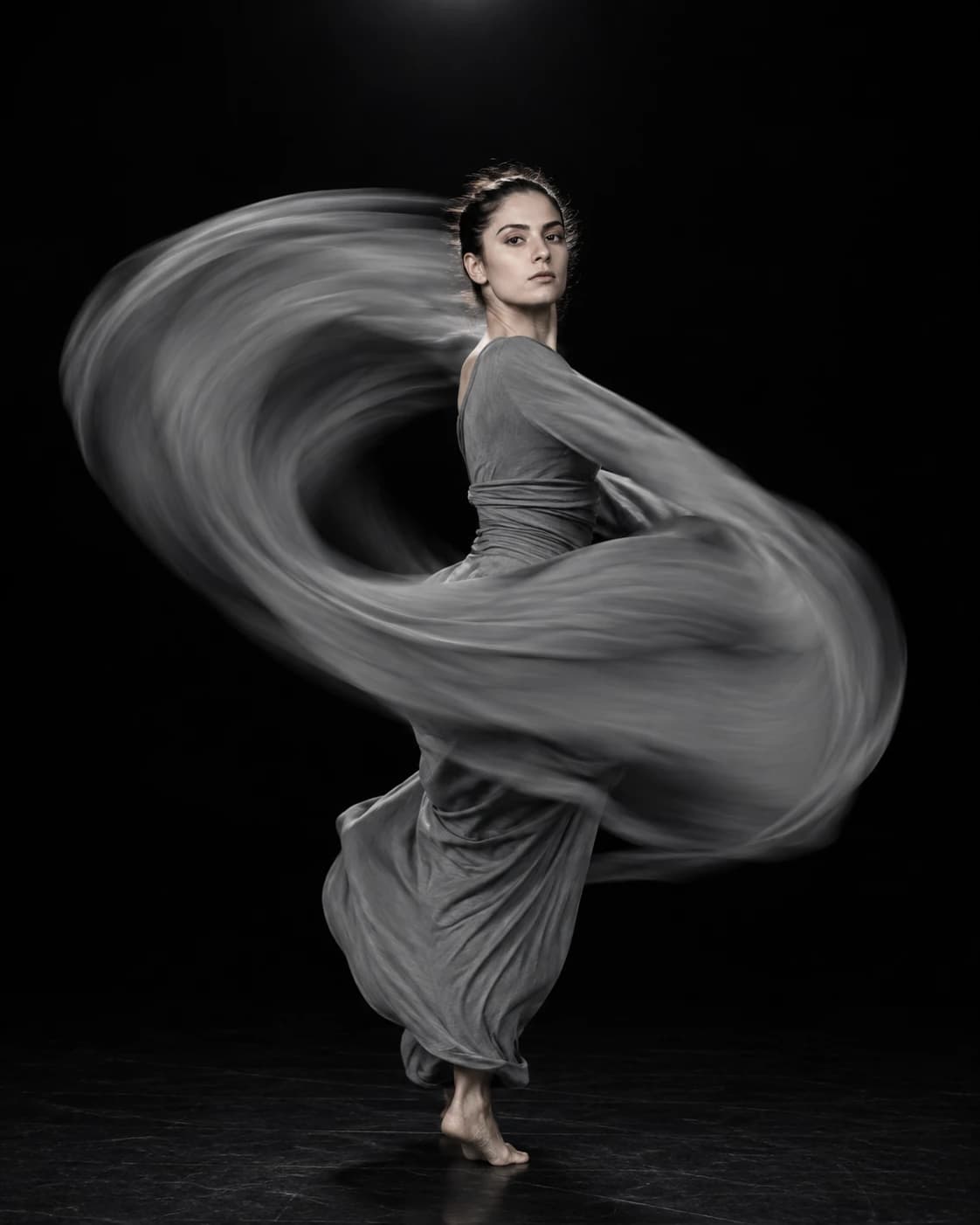 AI fine-art photograph of a dancer mid-turn in a dark studio, her grey dress blurred into a sweeping arc.