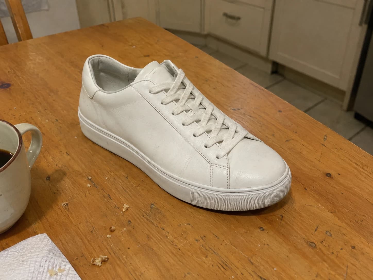 Phone-style snapshot of a white leather sneaker on a wooden kitchen table beside a coffee mug — the rough input before the finished image.